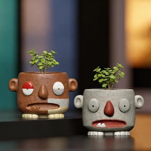 May include: Two ceramic planters shaped like stylized heads with ears and small feet. The left planter is brown with a red and white eye, while the right planter is gray with a red nose and mouth containing two white teeth. Both hold small green plants.