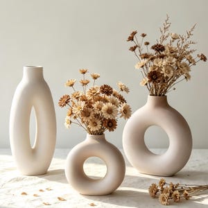May include: Three matte off-white ceramic vases of varying shapes. One tall oval vase, one small round donut-shaped vase, and one large round donut-shaped vase. The two round vases contain arrangements of dried brown and cream-colored daisy flowers.