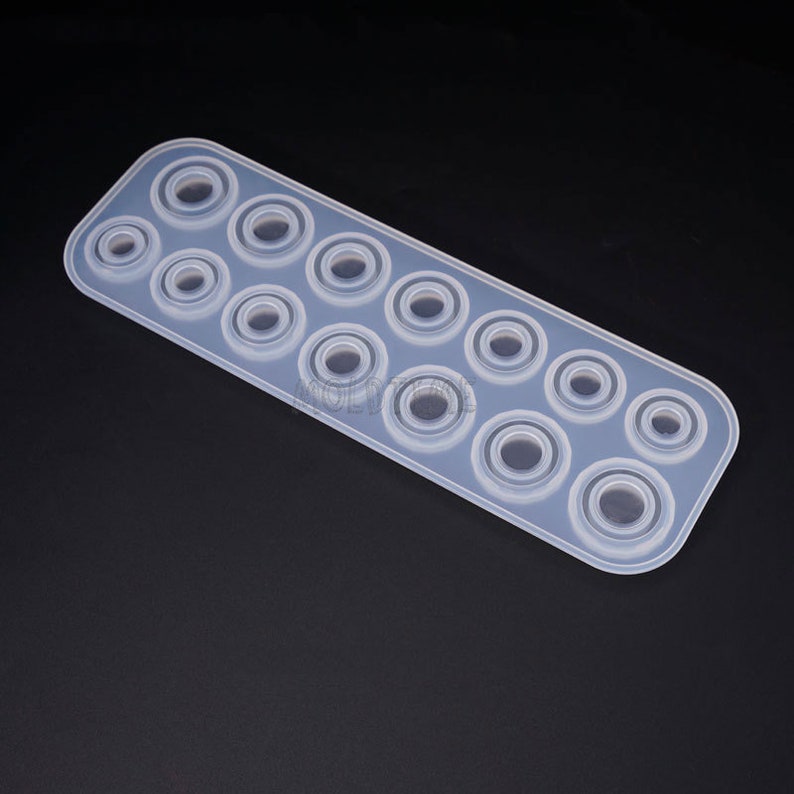 May include: A clear, rectangular silicone mold with multiple circular cavities of varying sizes. The mold is designed for crafting, likely for resin or similar materials, and is set against a dark background.