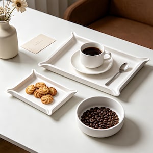 Puede incluir: Un juego de vajilla de cerámica blanca que incluye dos bandejas rectangulares con esquinas elevadas, un cuenco redondo lleno de granos de café tostados, una taza de café sobre un plato y una cuchara de plata, dispuestos sobre una superficie blanca.