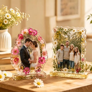 Peut inclure: Deux cadres photo décoratifs avec des accents floraux. L'un présente un couple de mariés, l'autre un portrait de famille. Les deux cadres sont transparents avec des fleurs incrustées et reposent sur une surface en bois.
