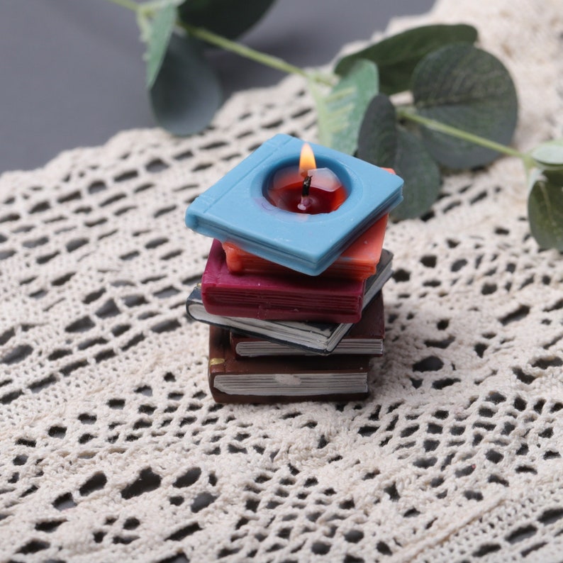 May include: A stack of book-shaped candles in various colors, including blue, red, and brown. The top candle is lit, with a flame. The candles are arranged on a white lace surface.