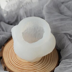May include: A translucent, white silicone mold for crafting, featuring a rounded, organic shape with a central cavity. The mold sits on a natural wood slice, with a soft, gray fabric in the background.