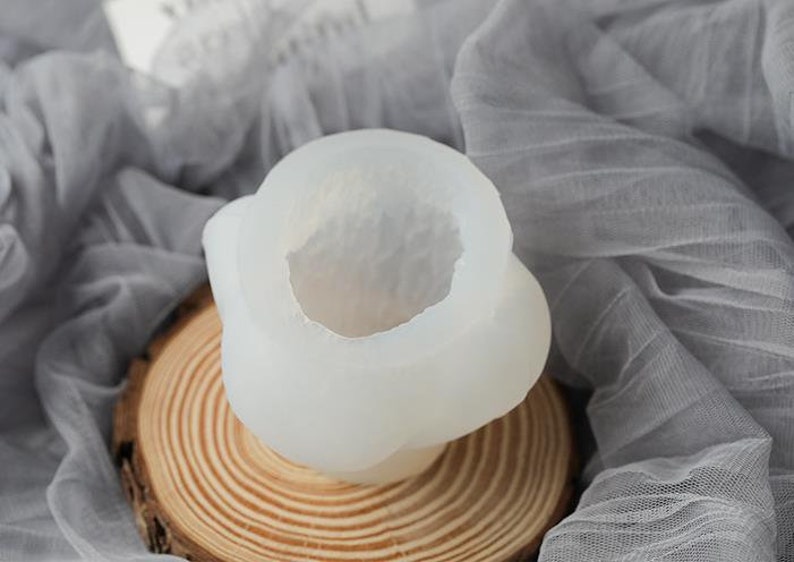 May include: A translucent, white silicone mold for crafting, featuring a rounded, organic shape with a central cavity. The mold sits on a natural wood slice, with a soft, gray fabric in the background.