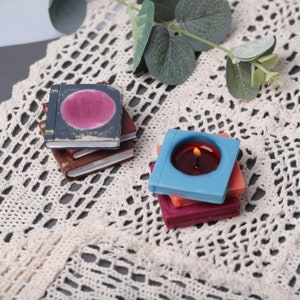 May include: A stack of miniature book-shaped candles in various colors, including blue, red, and brown. One candle is lit, with a small flame visible. The candles are arranged on a cream-colored, open-weave surface.