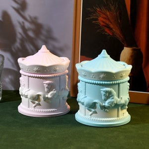 Op de afbeelding: Twee keramische containers met carrouselthema, één lichtroze en één lichtblauw, staan ​​naast elkaar. Elk heeft een gewelfd deksel en is versierd met carrouselpaarden. De containers staan ​​op een groene ondergrond.