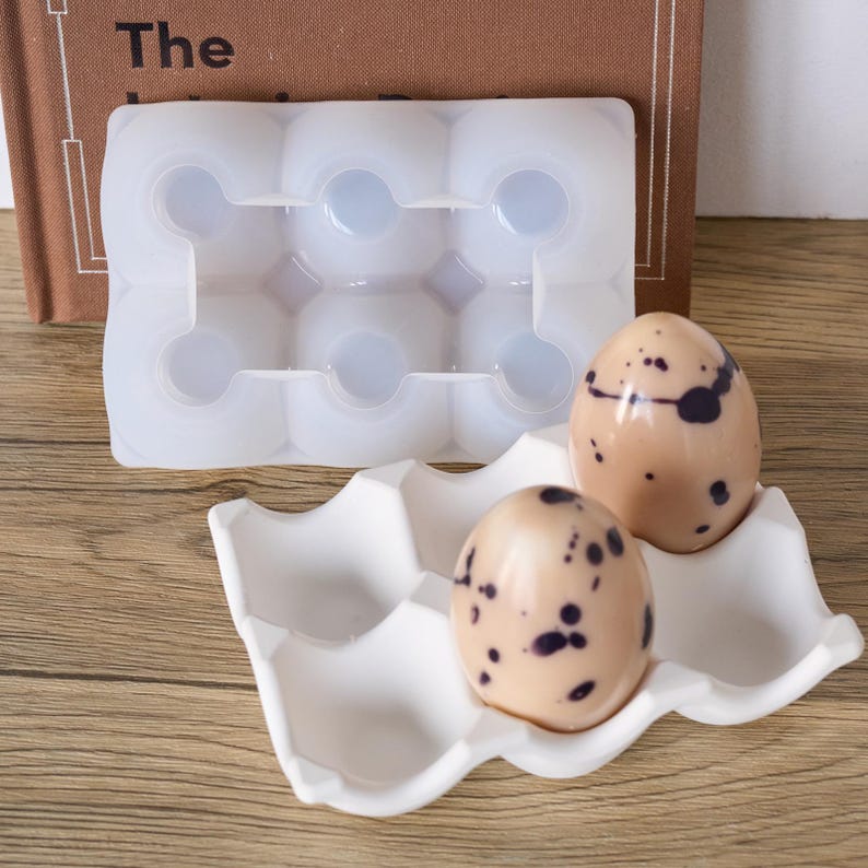 May include: A white silicone mould for creating six egg-shaped items, beside a white ceramic egg holder with two speckled eggs. The eggs are light brown with dark purple spots. The word "The" is visible on a book in the background.