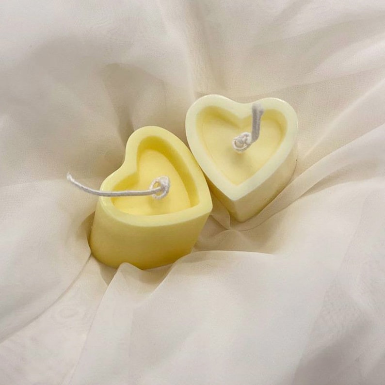 May include: Two heart-shaped candles with a pale yellow hue. Each candle has a white wick extending from the center. The candles are resting on a white, textured fabric, creating a soft and romantic aesthetic. The candles are a charming decorative item.