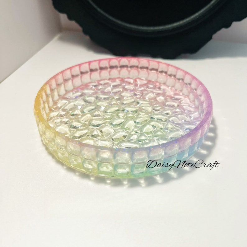 May include: A round, iridescent resin tray with a textured surface and a raised, square-patterned edge. The tray transitions through a spectrum of colors, including yellow, pink, and blue. The text "Daisy NoteCraft" is visible on the tray.