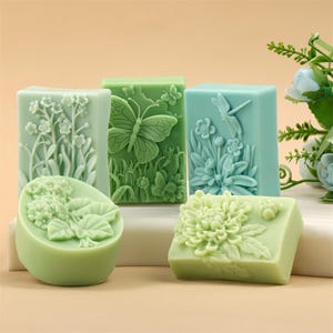 Puede incluir: Un surtido de jabones verdes y azules con diseños florales e insectos. Los jabones presentan tallas detalladas de mariposas, flores y una libélula. Los jabones están dispuestos sobre una superficie blanca sobre un fondo neutro.