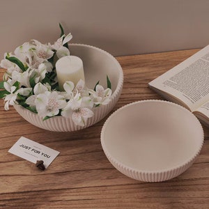 Puede incluir: Dos cuencos decorativos blanquecinos con diseño acanalado. Un cuenco contiene flores blancas y una vela. Una tarjeta con el texto "JUST FOR YOU" es visible. Los cuencos están sobre una superficie de madera.