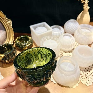 May include: A selection of decorative glass candle holders in different forms. The image shows a dark green, patterned bowl, alongside several white and clear candle holders. Some have textured surfaces, others are smooth.