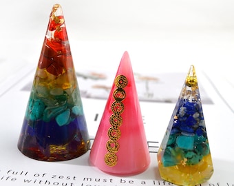 May include: Three colorful resin cone-shaped sculptures. One cone features a rainbow of stones, another is pink with gold symbols, and the third has blue and yellow stones. The cones are on a white surface.