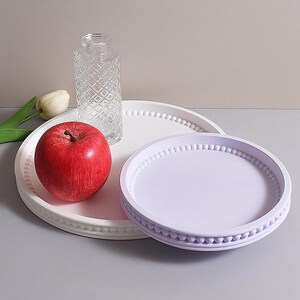 May include: A red apple, a clear glass vase, and two decorative trays. One tray is white with a beaded edge, and the other is lavender with a similar beaded design. A white tulip is visible.