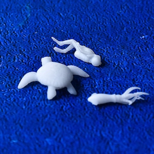 May include: White miniature sea creatures on a blue background. The image features a turtle, a diver, and a squid. The objects are small and appear to be made of a smooth material. The background is a textured blue.