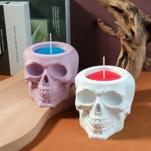 May include: Two skull-shaped candle holders. One is light pink with a blue candle, the other is white with a red candle. The candles have white wicks. The skull-shaped holders are on a wooden surface.
