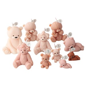 May include: A collection of teddy bear candles in various shades of pink and brown. The candles are in different poses, some sitting, some lying down, and some holding smaller bears. Each candle has a small tag attached.