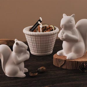 Peut inclure: Deux figurines d'écureuils en céramique blanche, l'une sur une base en bois, et un panier avec des fruits secs et des noix. Les écureuils sont en position assise, tenant quelque chose dans leurs pattes. Le panier porte une étiquette "HANDMADE".