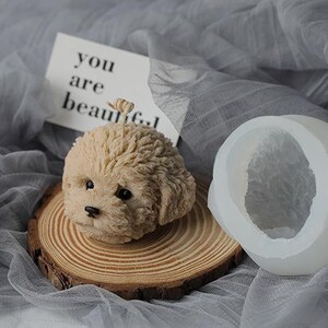 May include: A beige dog-shaped candle with black eyes and nose, resting on a wooden slice. Beside it is a white silicone mold in the shape of the dog's head. A card with the text "you are beautiful" is in the background.