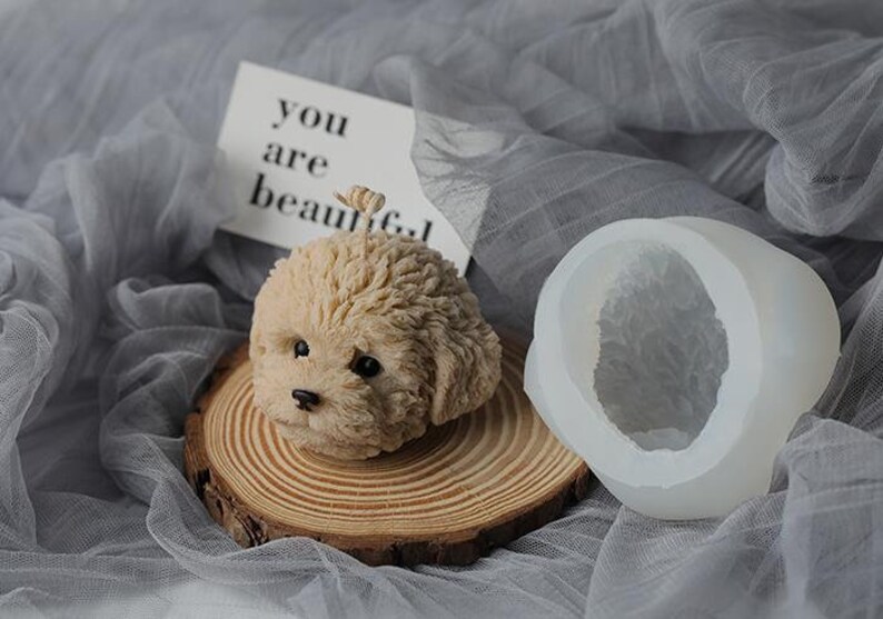 May include: A beige dog-shaped candle with black eyes and nose, resting on a wooden slice. Beside it is a white silicone mold in the shape of the dog's head. A card with the text "you are beautiful" is in the background.