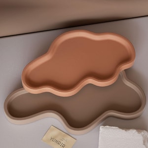 May include: Two cloud-shaped ceramic trays in earthy tones. The top tray is a warm terracotta color, while the bottom tray is a soft beige. A small card with the text "YUMRIR" is visible.