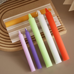 Może przedstawiać: Kolekcja kolorowych świec stożkowych w odcieniach pomarańczowego, białego, zielonego, fioletowego i różowego. Świece ułożone są na drewnianej powierzchni, prezentując swoje żywe odcienie i gładką fakturę. Idealne do dekoracji wnętrz.