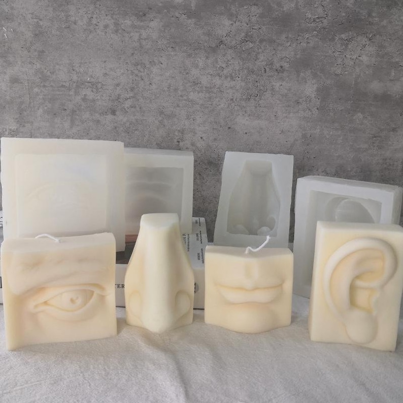 May include: A collection of cream-colored candles shaped like facial features, including an eye, nose, lips, and ear. The candles are arranged on a white surface, with molds in the background. The candles are unscented and have a minimalist aesthetic.