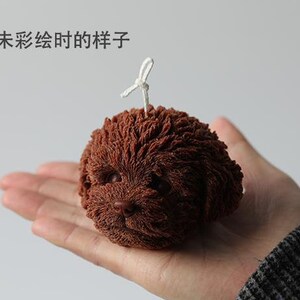 May include: A brown, dog-shaped candle with a textured surface. The candle has a white wick and the text "眼睛未彩绘时的样子" above it. The candle is held in a person's hand.