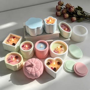 Pode incluir: Uma coleção de velas de várias formas em recipientes de tons pastel, incluindo formas quadradas, redondas, de coração e de abóbora. Várias velas estão acesas e apresentam pétalas de flores secas e pequenas pinhas sobre a cera.