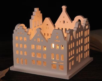 May include: A white ceramic candle holder shaped like a row of stylized houses. The houses have cut-out windows and doors, allowing light to shine through. The candle holder is on a dark surface, with a soft glow emanating from within.