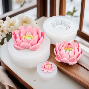 May include: A collection of pink lotus flower molds and finished flowers. The flowers have yellow and green centers. The molds are white and cylindrical. The finished flowers are displayed on a white surface and a wooden tray.