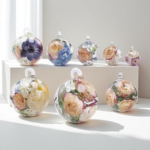 Pode incluir: Uma coleção de enfeites de vidro transparente, cada um preenchido com flores secas em várias cores. Os enfeites têm uma argola no topo para pendurar. As flores incluem rosas, hortênsias e margaridas.