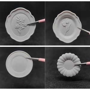 May include: Four small, white decorative plates with embossed floral and geometric designs. One plate features a floral design, another a tulip, a third has a beaded edge, and the last has a scalloped edge. A pink and silver tool points to each plate.