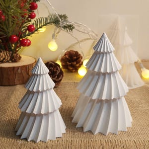 Puede incluir: Dos decoraciones blancas de árbol de Navidad en capas. Los árboles tienen un diseño texturizado y en capas y están colocados sobre una superficie de arpillera. Luces decorativas y piñas en el fondo sugieren un tema festivo.