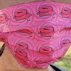 May include: A pink face mask with a repeating pattern of red lips dripping with pink liquid. The mask is made of fabric and has pleats.