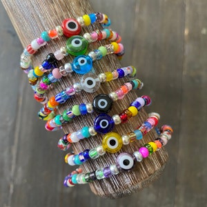 May include: A stack of colorful beaded bracelets with evil eye charms. Each bracelet features a different color combination and a unique evil eye charm.