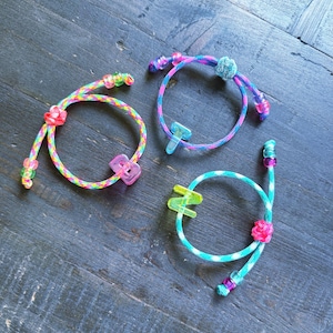 May include: Three colorful handmade bracelets. One is rainbow-colored, another is pink and blue, and the third is turquoise and white. Each bracelet features a unique clasp and decorative beads. The bracelets are displayed on a dark wooden surface.