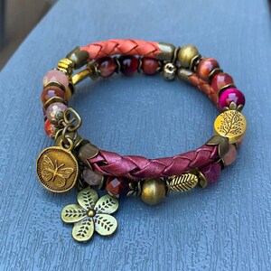 May include: A multi-strand bracelet featuring braided coral and deep pink leather cords, interspersed with faceted beads in shades of red, pink, and orange. It includes antique brass-toned metal charms of a butterfly and a flower.