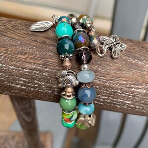 May include: A multi-strand beaded bracelet featuring a variety of round beads in shades of turquoise, green, blue, and dark purple. The bracelet includes silver-toned metal charms shaped like a leaf, a rabbit, and a butterfly.