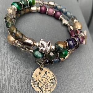 May include: A multi-strand wrap bracelet featuring braided leather, faceted green and purple beads, and metallic accents. A round brass-toned charm hangs from the bracelet, engraved with a detailed illustration of a fawn among plants and stars.
