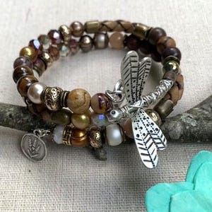 May include: A multi-strand bracelet with a dragonfly charm. The bracelet features a mix of brown, gold, and pearl beads, with a braided leather strand. A silver dragonfly charm and a small silver charm with a floral design add detail.
