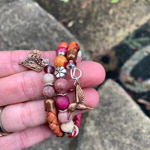 May include: A multi-strand bracelet with a variety of beads in shades of pink, orange, and brown. The bracelet features a copper-colored hummingbird charm and a heart-shaped charm. The beads are made of various materials, including stone and wood.