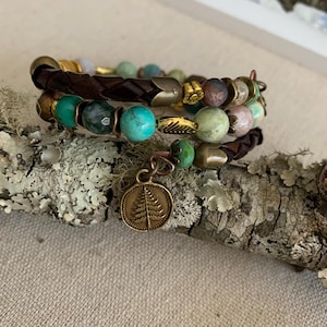 May include: Three stacked bracelets with a nature-inspired theme. The bracelets feature a mix of turquoise, green, and brown beads, along with a braided leather band and gold-colored accents. One bracelet has a charm with a tree design.