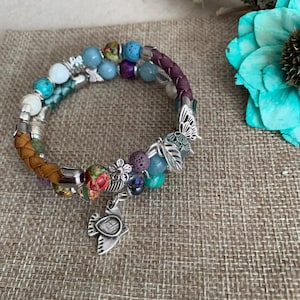 May include: A multi-strand bracelet featuring a braided purple leather band and colorful beads. The bracelet includes silver charms of a butterfly and a bird. The beads are in various colors, including turquoise, red, and purple.