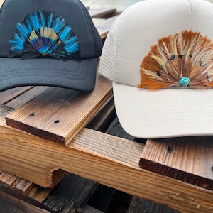 Puede incluir: Dos gorras de camionero con plumas. La gorra de la izquierda es negra con un dise&ntilde;o de plumas azules y negras. La gorra de la derecha es blanca con un dise&ntilde;o de plumas marrones y naranjas.