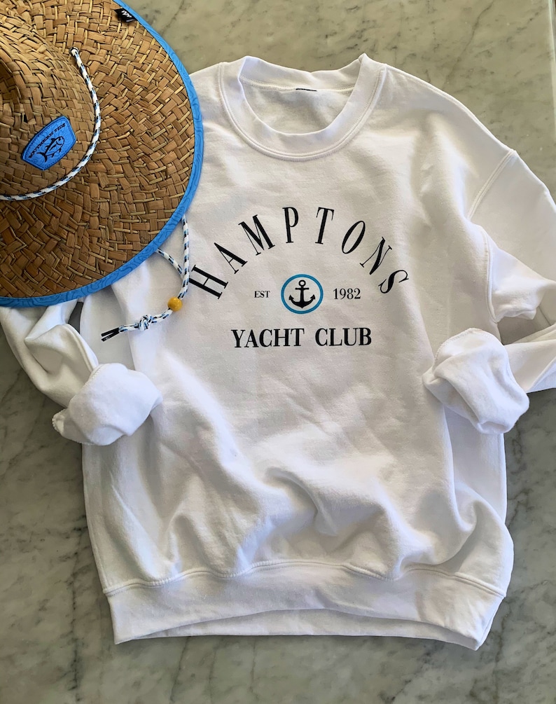 May include: White sweatshirt with a black graphic that says "Hamptons Yacht Club" with an anchor logo and the text "Est. 1982".