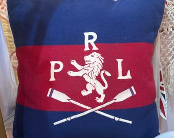 May include: A navy blue square pillow with a red stripe across the center. The pillow features the letters "RPL" above a white lion and crossed oars. The pillow is likely for home decor.