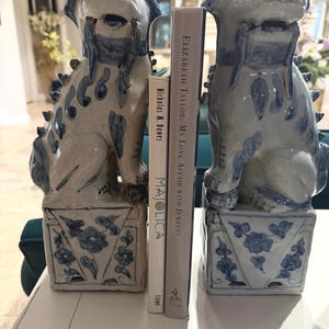 May include: Two white and blue ceramic foo dog statues with floral patterns on their bases, flanking two books. The books are titled "Majolica" by Nicholas M. Dawes and "Elizabeth Taylor: My Love Affair with Jewelry" by Elizabeth Taylor.