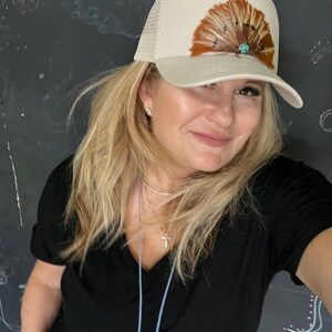 Puede incluir: Una mujer con cabello rubio lleva una gorra de camionero beige con un dise&ntilde;o de plumas marrones y un acento de piedra turquesa. Lleva una camiseta negra de cuello en V y un collar de plata.