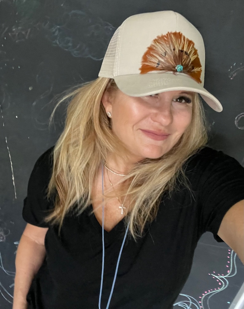 Puede incluir: Una mujer con cabello rubio lleva una gorra de camionero beige con un dise&ntilde;o de plumas marrones y un acento de piedra turquesa. Lleva una camiseta negra de cuello en V y un collar de plata.